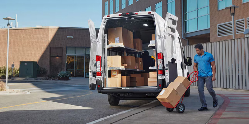 A person unloading packages from a white delivery van using a hand truck in an urban setting.